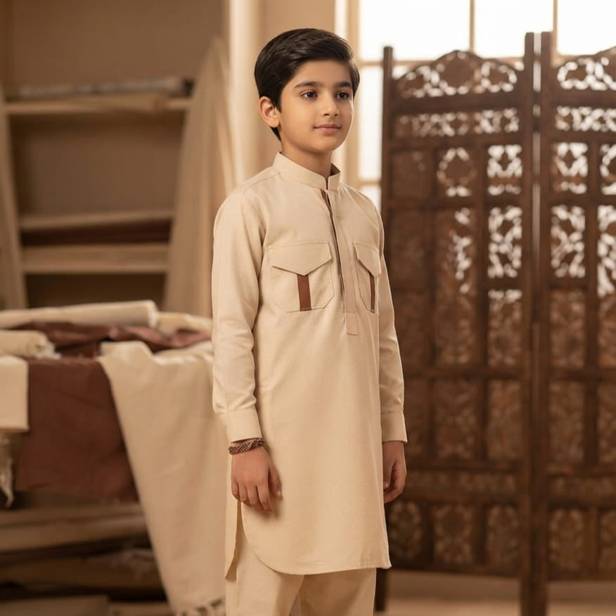 A young boy wearing a beige long-sleeve kurta and matching trousers, standing in a warm-toned room with a carved wooden screen behind him.